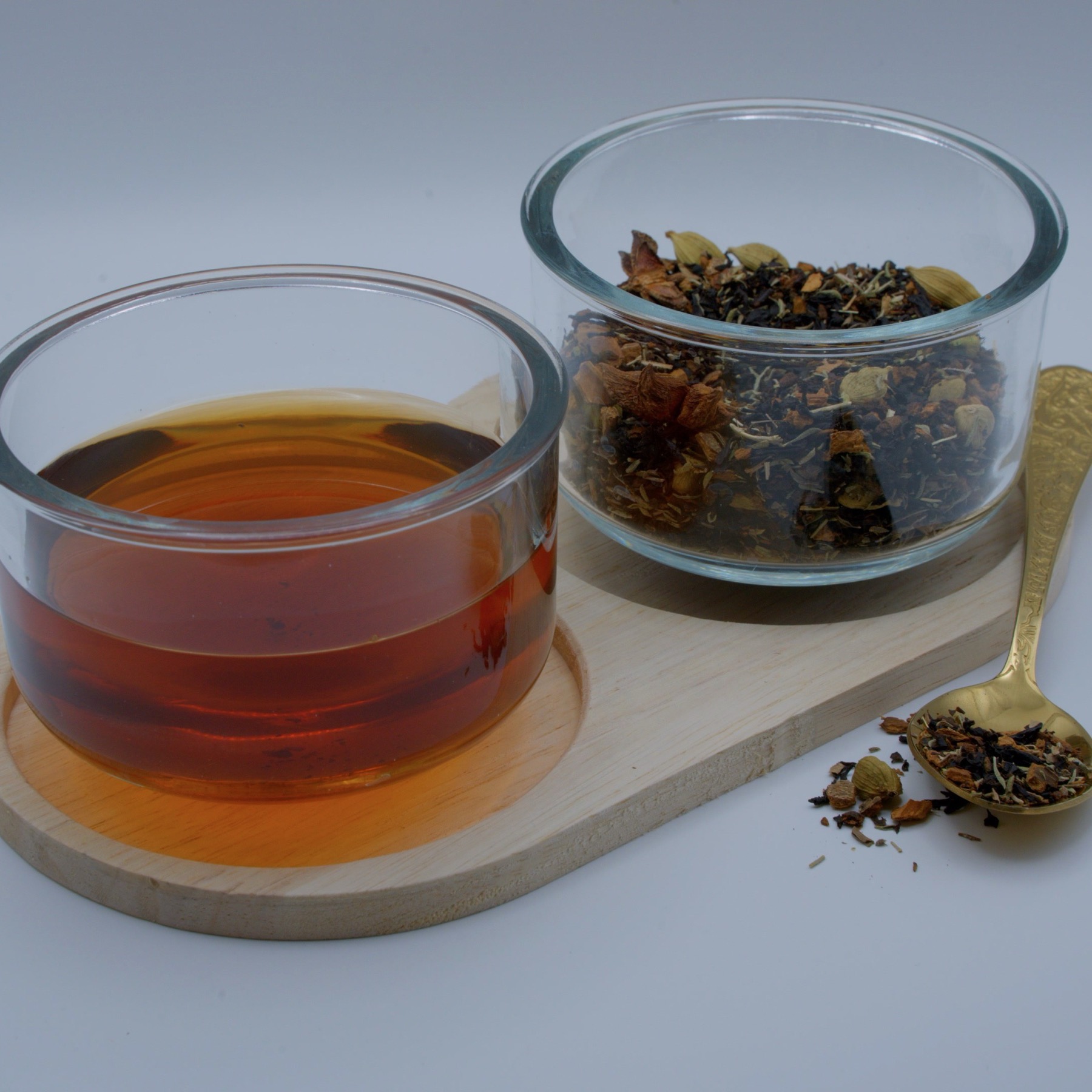 A glass bowl of brewed russet tea beside a bowl of black tea leaves with cardamom pods and cinnamon on a wooden board, with a gold spoon