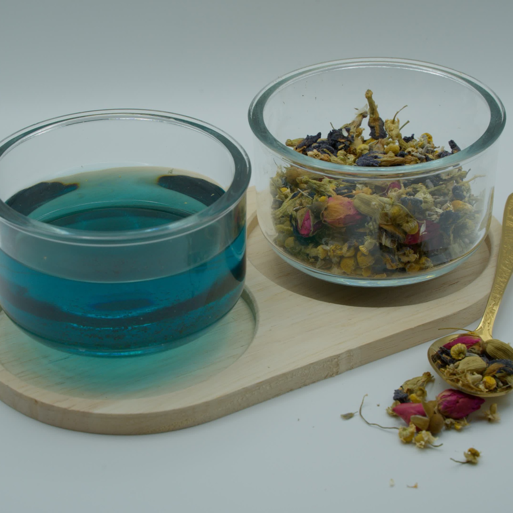 A glass bowl of brewed tea, turquoise from butterfly pea flower, beside a bowl of chamomile and rose buds on a wooden board