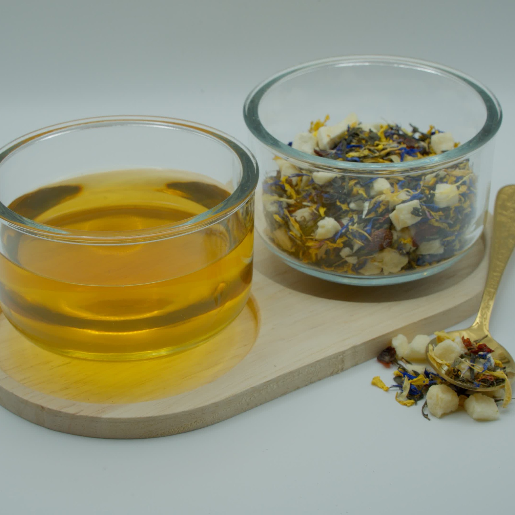 A glass bowl of brewed amber tea beside a bowl of green tea leaves, blue cornflower petals and dried peach on a wooden board
